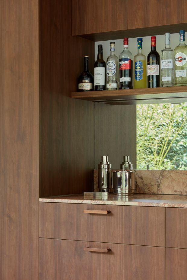 Close-up of a sophisticated built-in home bar, featuring dark timber cabinetry and shelving, a polished pinkish natural-stone benchtop and matching backsplash, chrome cocktail shakers, and a curated selection of liquor bottles. A window behind the counter provides greenery views and natural light. The cabinetry has simple contemporary slab fronts with timber handles.
