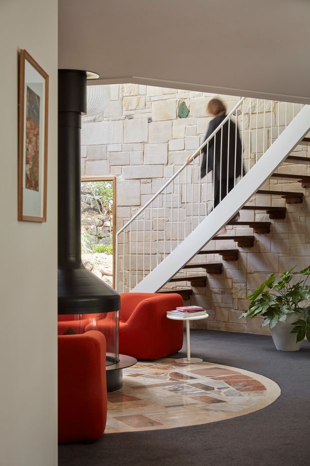 Interior view of a distinctive living and circulation area with an open-riser staircase, white metal balustrade, and exposed pale stone block wall. A black suspended fireplace, sculptural red lounge chairs, a small round side table, indoor planting, framed artwork, and a large irregular circular patterned stone or tile inlay define the space. Natural light enters through a small window and open stairwell area, creating a warm, eclectic modernist atmosphere.