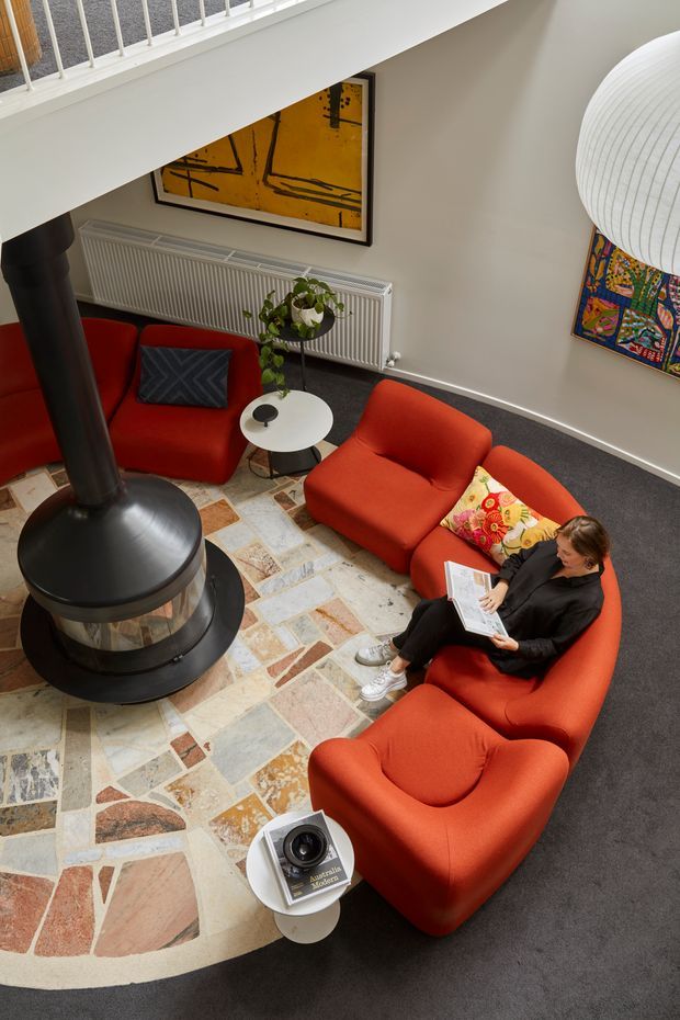 A double-height living room viewed from above, furnished with vivid red-orange upholstered lounge chairs and a curved sofa arranged around a freestanding black cylindrical fireplace. The space has a large irregular-patterned stone or tile floor rug, dark grey carpet, a white radiator, abstract wall art, a colorful textile wall hanging, a small round side table, and a white balustrade overlooking the room. A person is seated reading on the sofa.