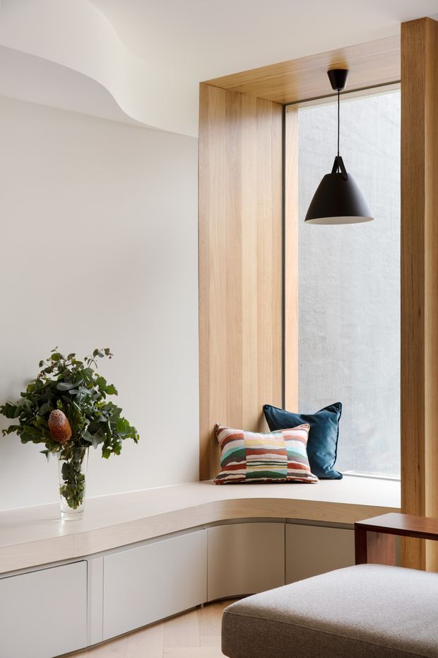 A refined, minimalist interior corner featuring a curved built-in window seat with pale cabinetry below, framed by warm timber reveals and a large vertical window. A black pendant light hangs over the nook, while layered cushions and a small indoor plant add restrained colour and texture. The foreground includes a softly upholstered bench or ottoman and a slim timber side table. The space is bright, calm, and carefully detailed, with a sculptural curved ceiling transition.
