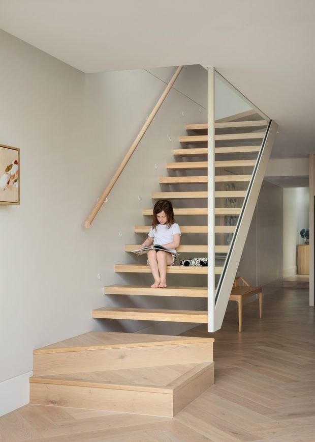 Interior view of a contemporary residential stairway with open-riser light timber stairs, a white structural stringer, glass balustrade, and timber handrail. The stair connects to a hallway and upper level, with light timber flooring in a herringbone pattern and neutral walls. A child is seated on the stairs.