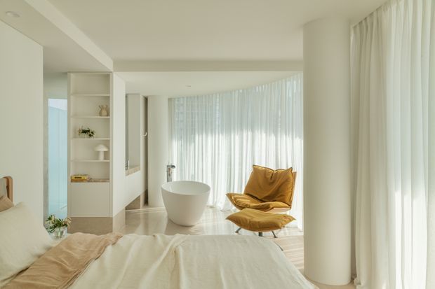 A bright, serene bedroom with a partially visible bed in the foreground, a freestanding white bathtub near the center, and a sculptural mustard lounge chair with matching ottoman beside expansive sheer-curtained glazing. The room features a restrained white and cream palette, integrated open shelving, smooth pale flooring, rounded columns, and abundant diffused daylight.