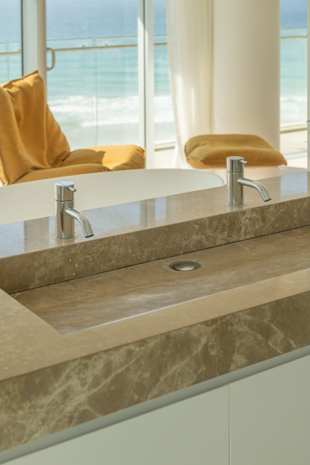 Close-up interior photograph of a refined bathroom vanity with a wide integrated stone basin, two chrome mixer taps, matching stone counter and backsplash, pale built-in cabinetry, and a large mirror. A bright ocean view and mustard lounge chairs are visible through the glazing and reflected in the mirror.