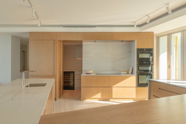 A bright contemporary kitchen with extensive light timber cabinetry, a large pale stone island benchtop, integrated black appliances, and a full-height pale stone splashback. Track lighting, streamlined joinery, and large glazed doors create a clean, open, sunlit interior.
