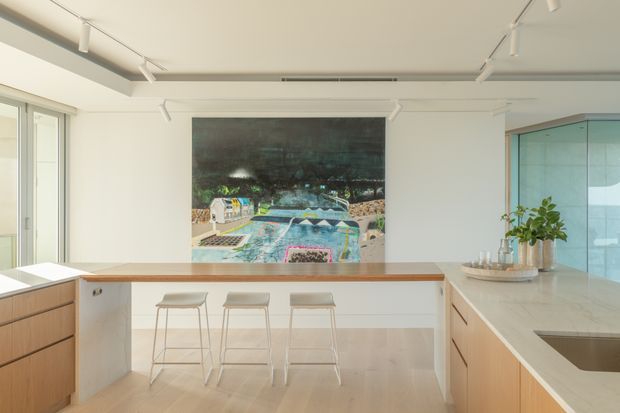 Bright, open-plan contemporary kitchen with a long peninsula and three white bar stools, pale timber cabinetry, a light stone benchtop, white walls, track lighting, and a large artwork on the rear wall. Full-height glazing and a glass-enclosed area are visible at the sides, with indoor plants and styling on the benchtop.