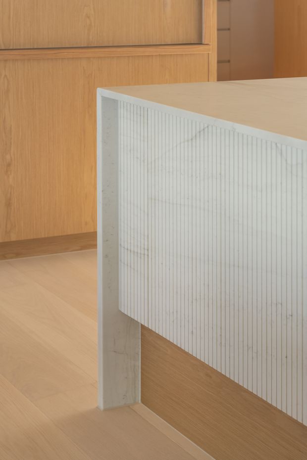 Close-up interior detail of a minimalist built-in island or counter with a pale stone or concrete top and vertically ribbed white panelled side. Warm timber cabinetry, wall panelling, and timber flooring surround the feature, creating a refined, tactile kitchen or joinery-focused setting.