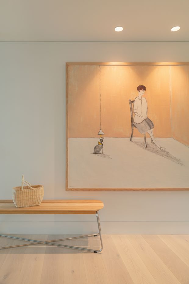A softly lit minimalist interior vignette featuring a low mid-century console table with a light timber top, slender metal frame and glass lower shelf. A woven basket sits on the console, while a large framed artwork with warm peach and neutral tones is mounted above. The room has pale warm-gray walls, recessed ceiling lights, and light timber flooring.