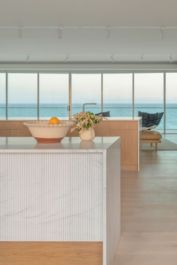 A bright, refined coastal kitchen with a large island featuring a vertically fluted pale stone face and warm timber base. The island has a light stone benchtop, while timber cabinetry extends behind it. Full-height windows provide expansive ocean views, and a sculptural bowl, fruit and floral arrangement decorate the island. A contemporary lounge chair is visible beyond the kitchen, with a pale timber floor and ceiling-mounted track lighting.