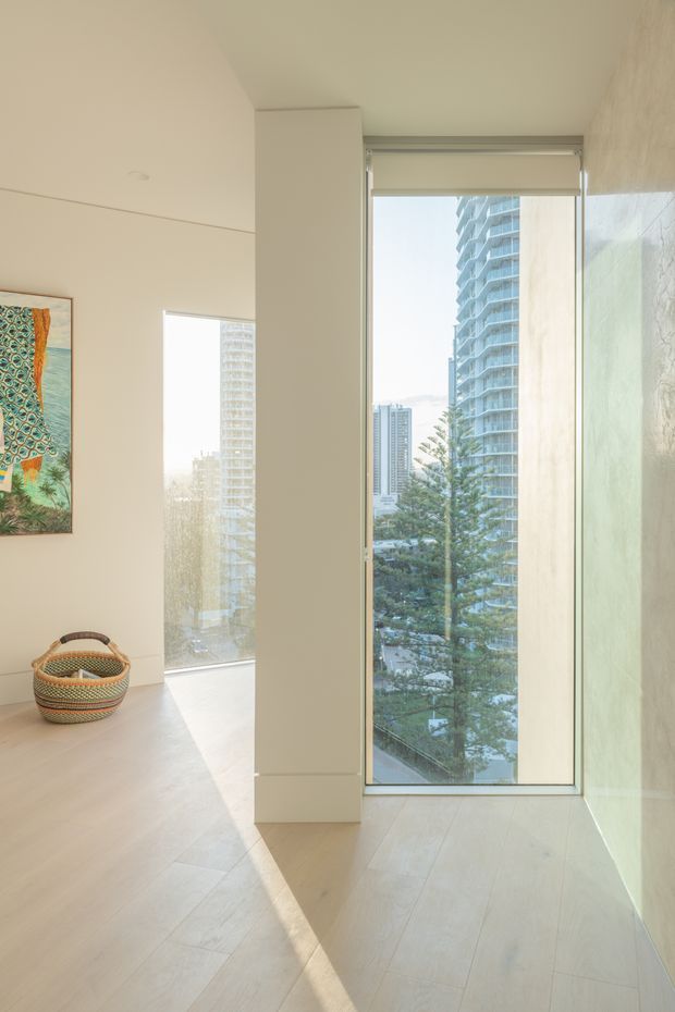 A bright, minimalist apartment interior with pale timber flooring, smooth off-white walls, and expansive floor-to-ceiling glazing overlooking high-rise buildings and trees. A narrow structural or joinery column divides the windows, while a colorful framed artwork and woven basket add subtle decorative detail. Strong sunlight creates long shadows across the floor.