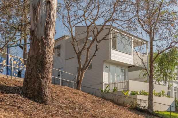 Exterior view of a contemporary multi-level residential building on a sloped, tree-lined site. The light-coloured building has horizontal weatherboard cladding, large glazed openings, projecting upper-floor volumes, balconies or terraces with simple metal and glass-like railings, and a partially visible colourful patterned feature wall at left.