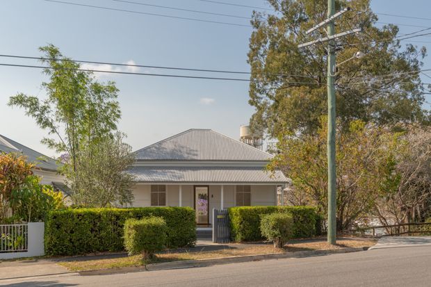 Street-facing view of a modest single-storey house with a symmetrical corrugated metal hipped roof, covered front verandah, pale rendered walls, and a central entry. Trimmed hedges, a low fence, mature trees, overhead utility wires, and a roadside verge frame the suburban residential facade.
