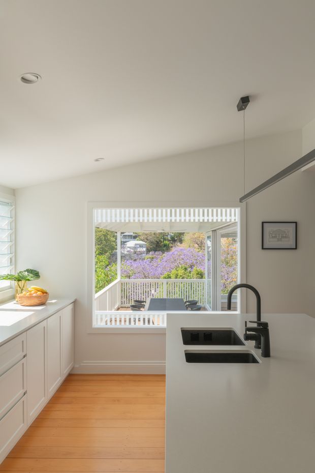 Bright contemporary kitchen interior with a large white island, integrated double sink and dark curved mixer tap, white perimeter cabinetry, warm timber flooring, and a large window opening to a leafy outdoor deck and garden. Minimal pendant lighting and restrained neutral finishes create an airy, refined space.