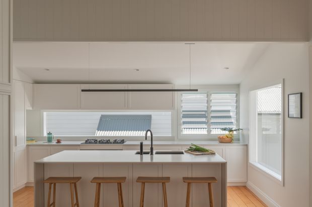 Bright contemporary kitchen with a large central island and four timber stools. The space features white built-in cabinetry, a light benchtop, integrated cooktop, double sink with dark tapware, horizontal louvre windows, timber flooring, and a slim linear pendant light. A glazed door and additional window provide views to the exterior.