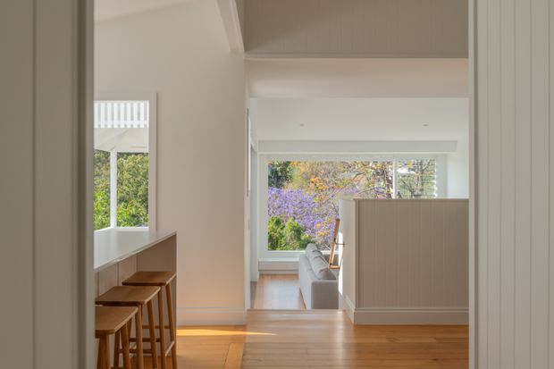 A bright, refined residential interior viewed from a kitchen or dining transition area toward a living room. A timber island or counter with several wooden stools occupies the left foreground, while a partially screened living area with a pale sofa is visible beyond. Large windows bring in abundant natural light and overlook leafy garden vegetation. The space features warm timber flooring, pale walls, vertical fluted joinery, and a high sloped or vaulted ceiling.