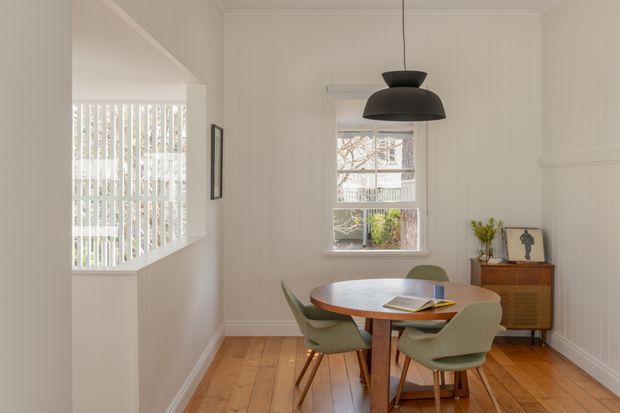A bright, compact dining room with a round timber dining table and four upholstered mid-century-style chairs. The room features warm timber flooring, white vertical wall panelling, simple white trim, a large window with vertical blinds, and a black sculptural pendant light. A timber sideboard with minimal decor sits against the far wall.