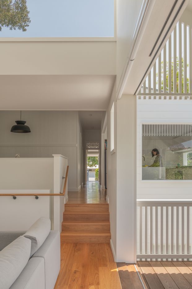 Bright contemporary multi-level residential interior showing a stairway and landing beside a double-height void. Warm timber flooring and stairs contrast with white painted walls, vertical balustrades, and large windows. A partial white sofa is visible in the foreground, with a hallway extending toward the rear and a glazed internal opening overlooking another area of the home.