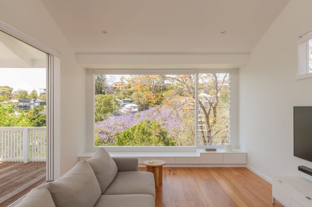 Bright contemporary living room with warm timber flooring, an L-shaped neutral sofa, a small round timber coffee table, low built-in media cabinetry, and a wall-mounted television. Large windows overlook leafy suburban gardens, while folding glass doors open onto a balcony or terrace. The room has a simple white-painted interior, recessed ceiling lighting, and abundant natural light.