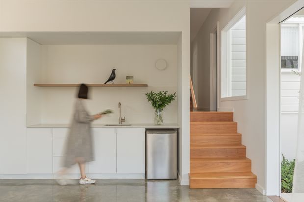 Bright, minimalist open-plan interior featuring a compact white kitchen with streamlined cabinetry, integrated sink and tap, timber floating shelf, and stainless-steel under-bench appliance. A timber staircase rises beside the kitchen, with large windows and a glazed door providing natural light. Polished grey concrete flooring and warm white walls create a clean, calm aesthetic.