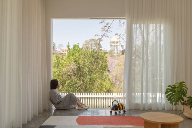 Bright, serene interior with floor-to-ceiling sheer curtains framing a large window and leafy suburban outlook. A person sits on a polished concrete floor beside a colourful rug and small decorative objects, with a round timber coffee table and indoor plant in the foreground. The space has a calm, minimalist feel and abundant natural light.