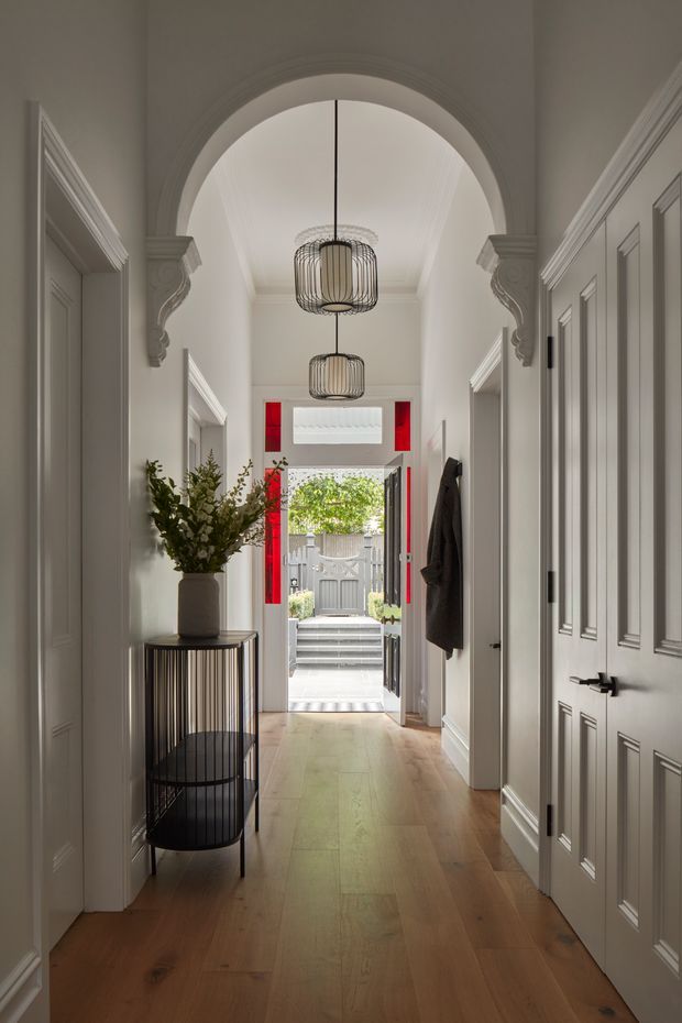 A refined residential hallway viewed toward an open front door. The corridor has warm timber flooring, pale grey panelled doors and walls, ornate architraves and corbels framing a barrel-vaulted ceiling, and two suspended ribbed pendant lights. A round black metal console table with a potted leafy arrangement sits along the left wall. Bright red door reveals and sidelights provide a bold accent, with steps and a small exterior courtyard visible beyond.