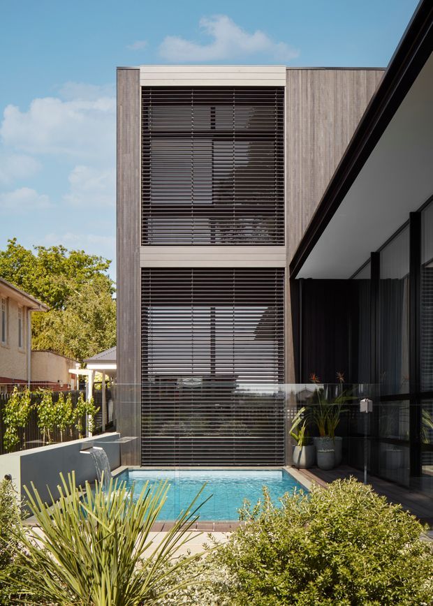 Exterior view of a contemporary multi-storey residence centred on a tall, vertically proportioned facade with extensive dark horizontal louvres screening the windows. A glazed, covered outdoor living area projects on the right, while a compact swimming pool, paving and landscaped planting occupy the foreground. The facade combines vertical timber-look cladding with pale horizontal trims and dark glazing.