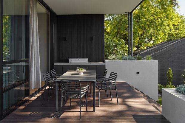 Contemporary outdoor dining terrace with a dark timber deck, black-framed glazing, and a covered outdoor kitchen or barbecue zone. A rectangular dining table with black metal chairs occupies the centre, while raised white rendered planters and greenery define the perimeter. The space overlooks a neighbouring dark-clad roof and leafy trees.