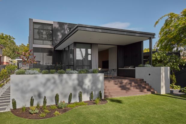 Exterior view of a contemporary multi-level residence with a prominent flat-roofed upper volume, dark vertical cladding, extensive glazing, and a sheltered entry or outdoor living area. A broad staircase leads through landscaped front garden beds to the covered porch, with rendered boundary walls and manicured lawn.