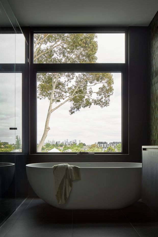 A moody, sophisticated bathroom featuring a freestanding oval bathtub positioned beneath large black-framed windows. The room has dark tiled flooring and walls, a textured green tile feature wall, reflective cabinetry or glazing on the left, and a leafy urban view beyond the windows.