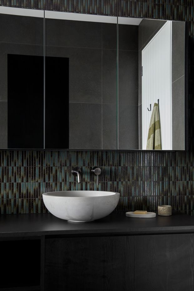 A dramatic, dark contemporary bathroom vanity featuring a white vessel basin on a black benchtop, black timber-look cabinetry, and a glossy vertically arranged iridescent dark tile splashback. A large mirror reflects an open white door with a hanging towel. Black and charcoal finishes create a high-contrast, moody atmosphere.