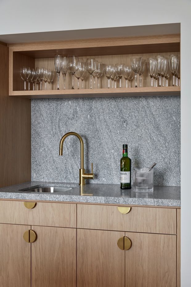 A compact, elegantly detailed bar or beverage station with warm wood cabinetry, a grey natural-stone benchtop and full-height splashback, brass gooseneck tap and hardware, and an undermount sink. An open upper shelf displays wine and cocktail glasses, while a bottle and tumbler sit on the counter. The design is refined and contemporary with a warm, tailored palette.