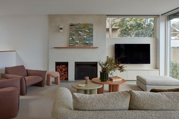 A refined contemporary living room with a warm neutral palette. The room features a textured plaster or concrete-look fireplace wall with two fireboxes, a timber mantel shelf, wall-mounted television, large corner windows, layered round timber coffee tables, and soft upholstered seating in muted mauve, cream, and taupe tones.