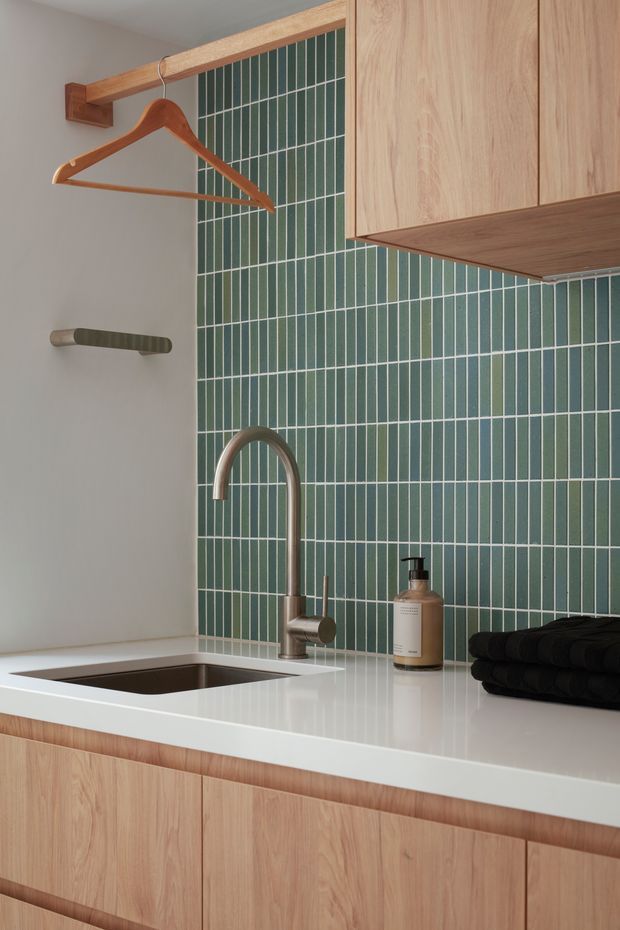 Close-up view of a compact contemporary kitchenette or wet-bar area. Pale timber cabinetry and a white benchtop surround a stainless-steel sink and brushed metal gooseneck tap. The backsplash is covered in vertically arranged narrow green ceramic tiles with light grout. A timber rail with a wooden hanger and a small wall-mounted metal rack are visible, along with a soap dispenser and folded dark towels.