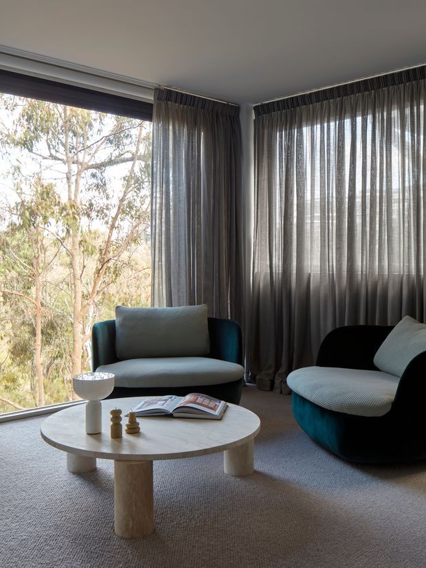 A refined living room corner with expansive corner glazing overlooking trees, layered dark sheer curtains, two sculptural upholstered lounge chairs, and a round light stone coffee table on a neutral carpet. The space has a calm, contemporary feel with strong natural light and dark teal and charcoal seating accents.