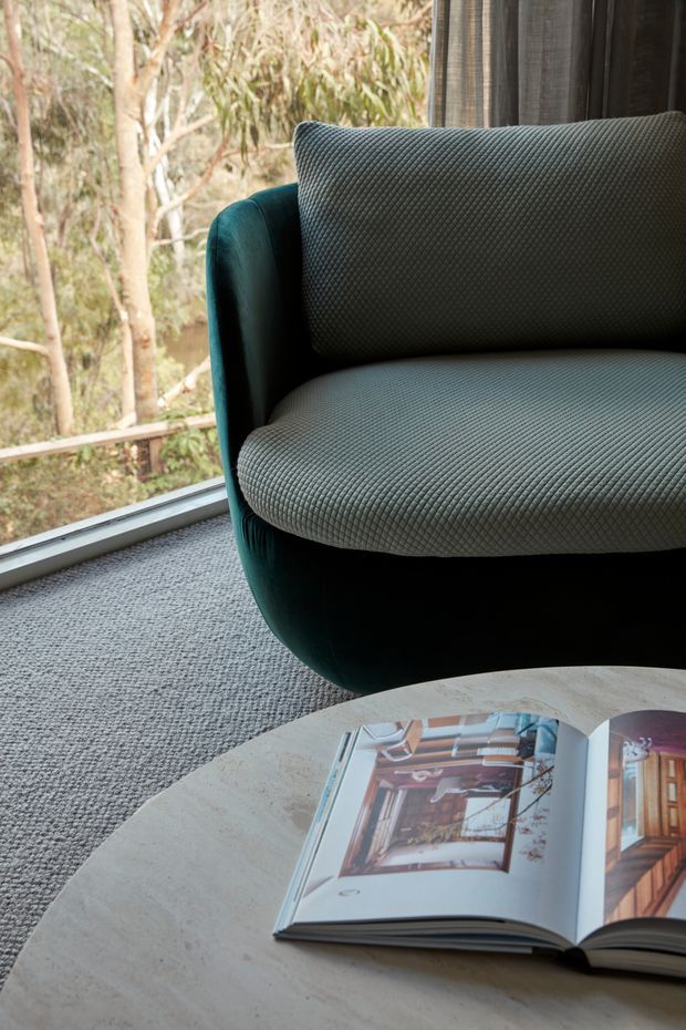 Close-up interior view of a refined seating area beside floor-to-ceiling glazing. A dark teal upholstered lounge chair with textured grey-green cushions sits on grey carpet, next to a round pale stone or concrete coffee table holding an open design book. Timber and leafy vegetation are visible outside through the window, while dark curtains frame the glazing. The scene has a calm, contemporary and nature-connected atmosphere.