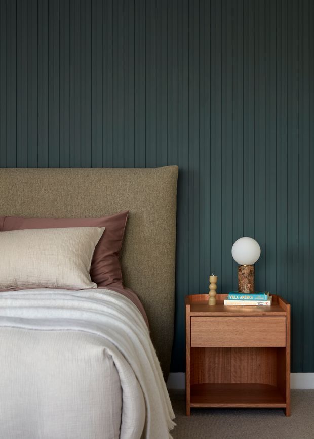 A close-up bedroom interior with a large upholstered bed and layered neutral bedding beside a compact mid-century-inspired timber bedside table. The wall is clad in dark vertical fluted or ribbed panelling, creating a moody backdrop. A spherical table lamp, small decorative object, and books sit on the bedside table. The palette combines deep blue-green, warm brown timber, taupe, blush, and cream.