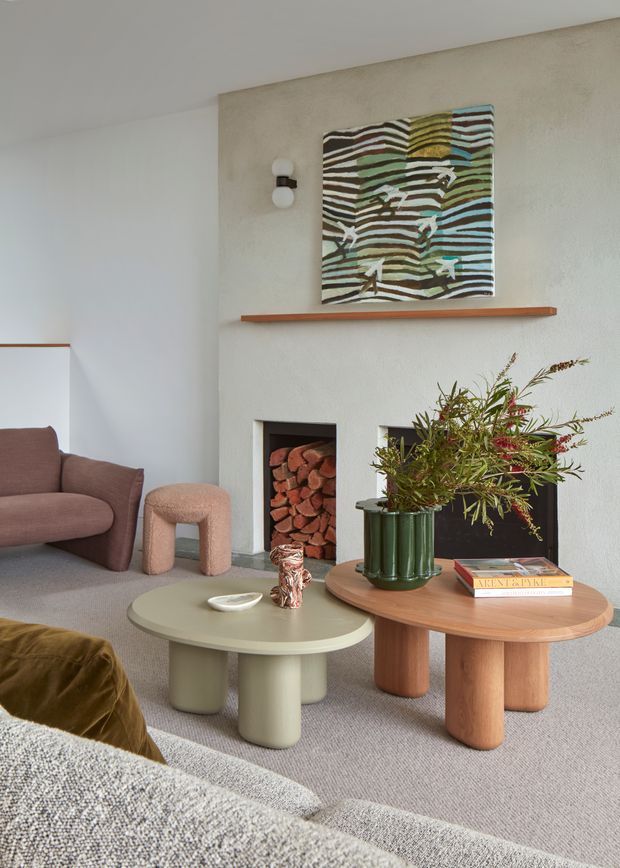 A warm, contemporary living room centred on a textured plaster fireplace wall with a wood mantel shelf and two recessed firewood niches. The room features rounded coffee tables, a curved upholstered armchair, a large neutral sectional sofa, a green ribbed vase with foliage, and abstract artwork above the fireplace. Soft natural tones and rounded furniture create a refined, inviting atmosphere.