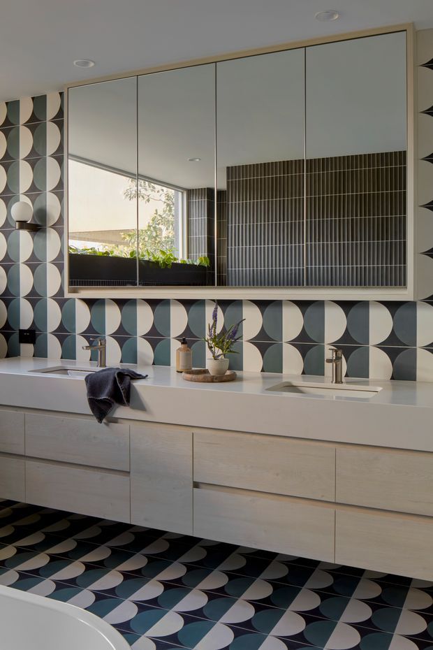 A stylish contemporary bathroom featuring a long floating light-wood vanity with two integrated white basins and chrome tapware. The walls and floor are covered in a bold geometric patterned tile made of repeating black, white, and muted blue-grey curved shapes. A large mirrored cabinet spans the vanity, reflecting a bright window, planter boxes, and vertically ribbed dark wall tiles. The room has recessed ceiling lighting and a clean, graphic, highly coordinated design.