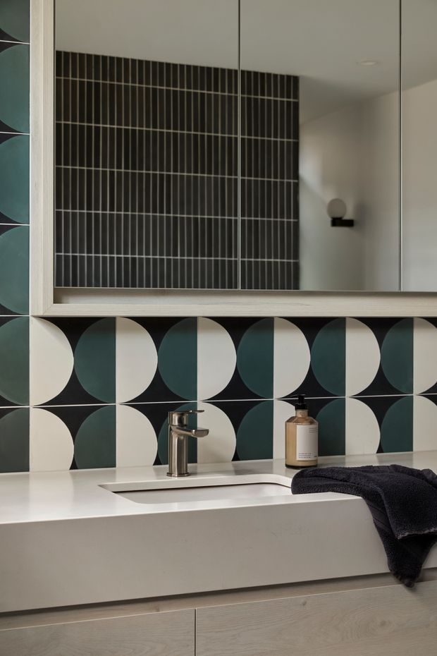 Close-up of a refined bathroom vanity featuring a white integrated countertop and undermount basin, brushed metal tapware, pale timber-look cabinetry, and a large framed mirror. The backsplash is a bold geometric tile pattern in dark teal, cream and black, with a dark vertically ribbed tile feature reflected in the mirror and a compact wall sconce.