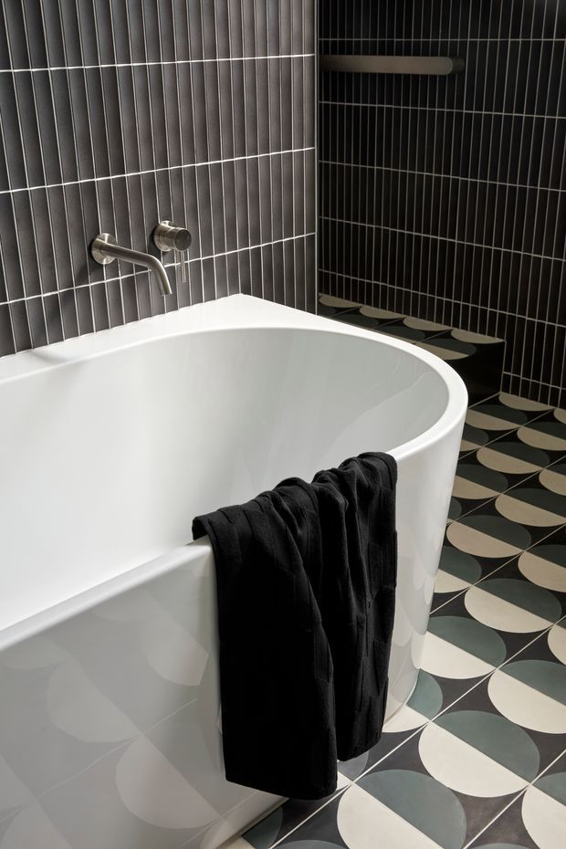 A dramatic contemporary bathroom featuring a freestanding white oval bathtub, wall-mounted brushed metal tapware, and dark vertically stacked rectangular wall tiles. The floor uses a bold geometric patterned tile in black, cream, and muted blue-grey, complemented by a black towel draped over the tub. A slim wall-mounted towel rail is visible in the shower or bath zone.