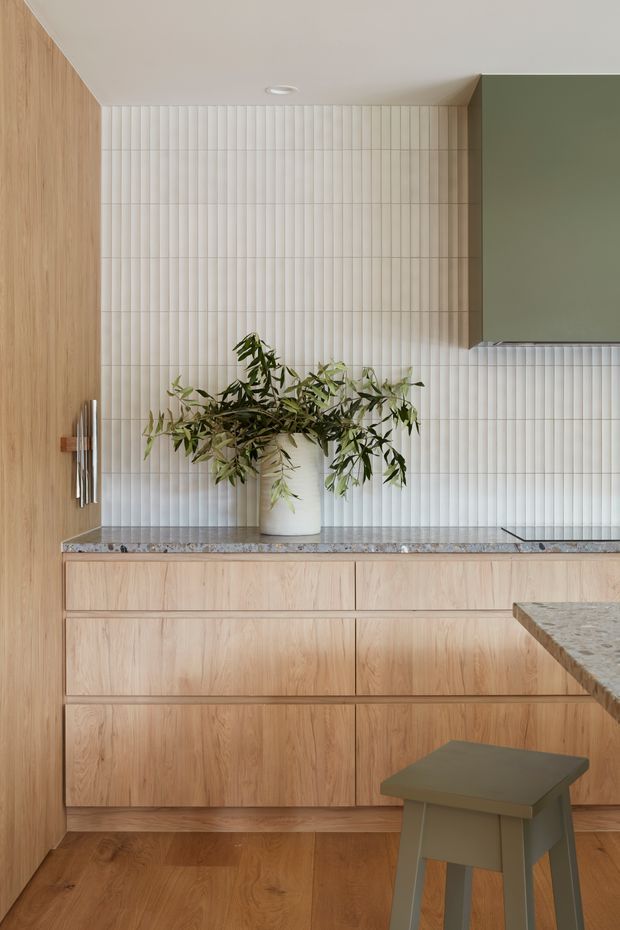 A refined contemporary kitchen vignette with full-height light timber cabinetry, a pale stone benchtop, vertically fluted white tile splashback, and a muted green upper cabinet. A small stool and leafy branch in a vase add warmth to the restrained composition.