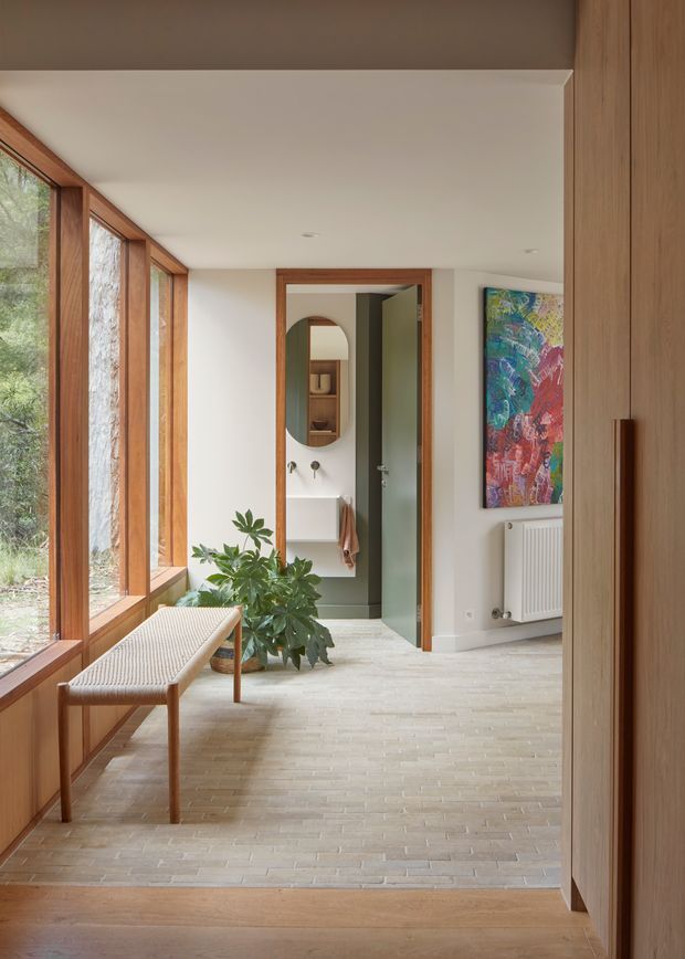 A refined residential hallway or entry passage with full-height timber-framed glazing along the left side, a built-in timber cupboard on the right, and a pale brick or stone floor transitioning to timber flooring in the foreground. A narrow upholstered timber bench sits beside the windows, while an open doorway reveals a compact powder room with a wall-mounted basin, oval mirror, and green-painted walls. The space is softly lit, warm, and contemporary, with a colourful artwork and indoor plant adding accents.