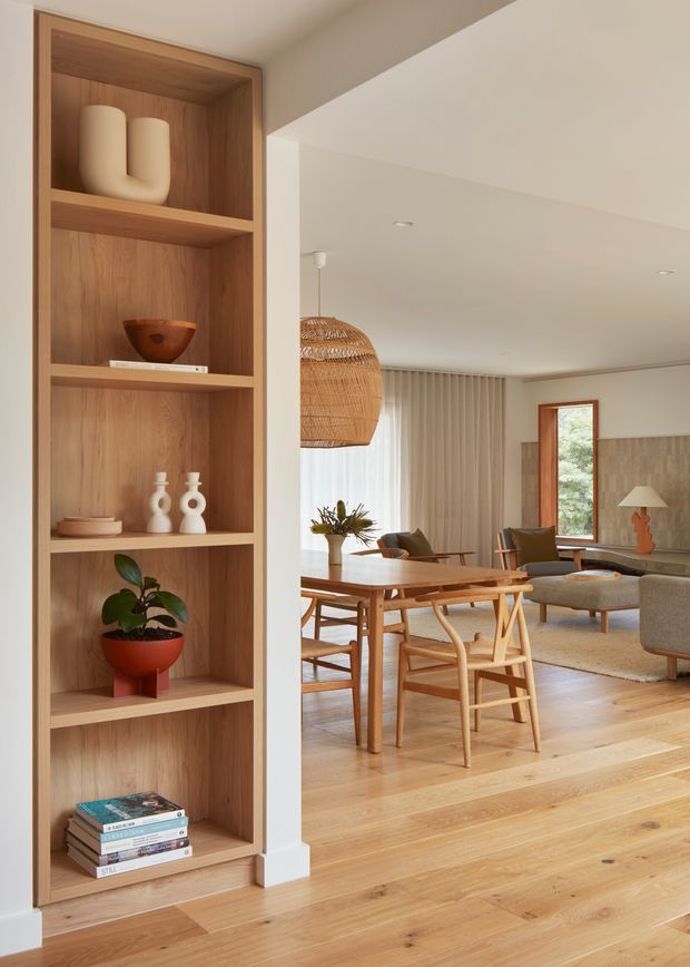 Warm, contemporary open-plan dining and living area viewed beside a full-height built-in timber shelving unit. The room features light timber flooring, a timber dining table and chairs, a woven pendant light, neutral upholstered seating, sheer curtains, recessed ceiling lights, and a stone or textured feature wall around a timber-framed window. The shelving displays ceramics, books, and a potted plant.