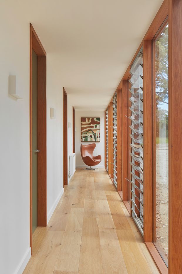 A long, bright residential hallway with pale timber flooring, white walls and ceiling, warm timber door frames, and a continuous wall of timber-framed louvred glazing on the right. Multiple doorways line the corridor, which terminates in a sculptural brown lounge chair and abstract artwork. The space has a clean mid-century-inspired character with strong natural light and a clear indoor-outdoor connection.