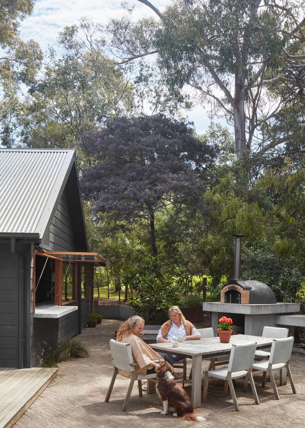 A landscaped outdoor entertaining area beside a dark weatherboard-clad building. The space features a paved dining terrace with a rectangular outdoor table and several chairs, a covered outdoor cooking structure with a domed masonry pizza oven and flue, raised garden beds, mature trees, and a small timber deck. Two people and a dog are seated at the table.