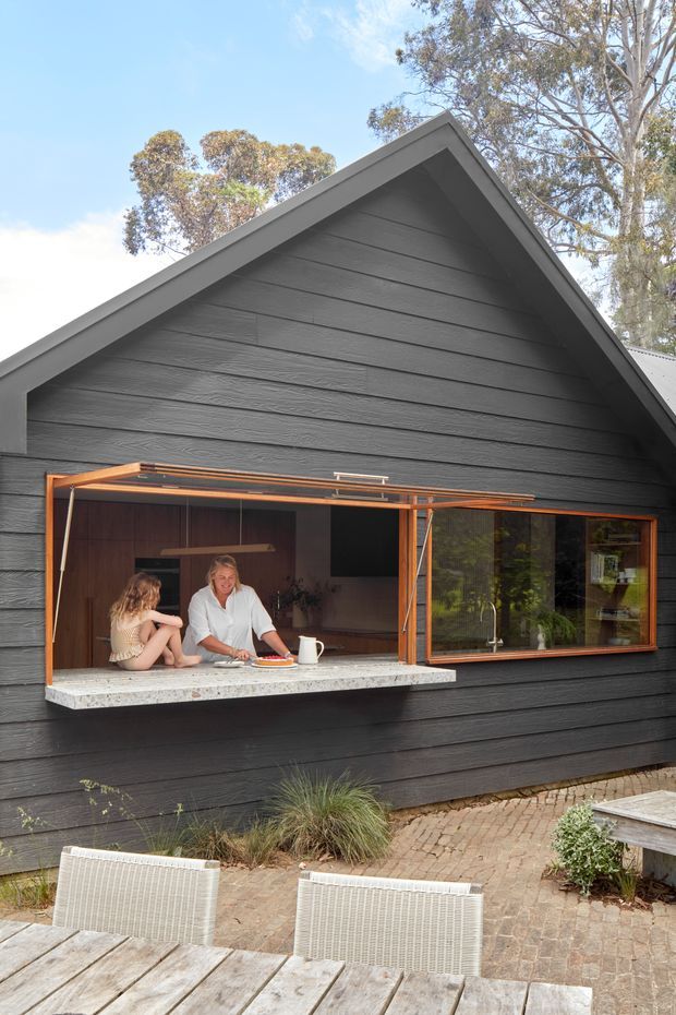 Exterior view of a dark charcoal, gable-roofed house with horizontal textured cladding and warm timber-framed glazing. A large bi-fold serving window opens onto a stone benchtop, connecting the interior kitchen or living area with a paved outdoor terrace. Timber decking, woven outdoor chairs, planting, and mature trees surround the structure.