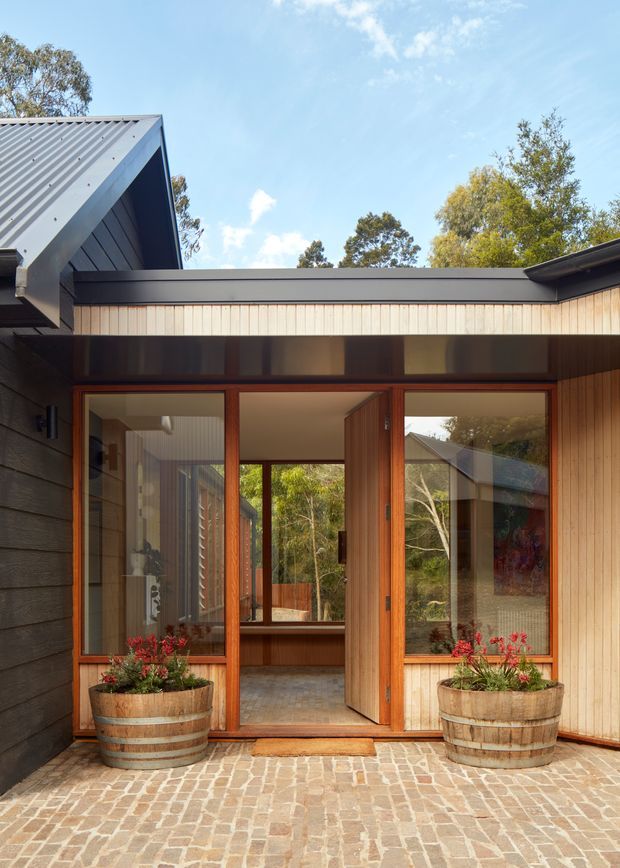 Exterior view of a contemporary timber-clad dwelling with a sheltered entry courtyard. The facade combines vertical pale timber boards, dark horizontal cladding, a pitched corrugated metal roof, and large timber-framed glazed doors, including an open central entry. The ground is paved in light irregular stone or brick, with two large timber planters flanking the doorway. Native trees are visible beyond the glazing and roofline.