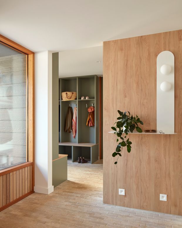 Warm contemporary entryway featuring a full-height light timber feature wall with an arched recessed niche and integrated display shelf, opening toward a built-in mudroom-style wardrobe. The space has pale timber flooring, white walls and ceiling, olive-grey joinery, and a timber-framed glazed opening with lower vertical timber panelling. Natural light and restrained styling create a calm, refined atmosphere.