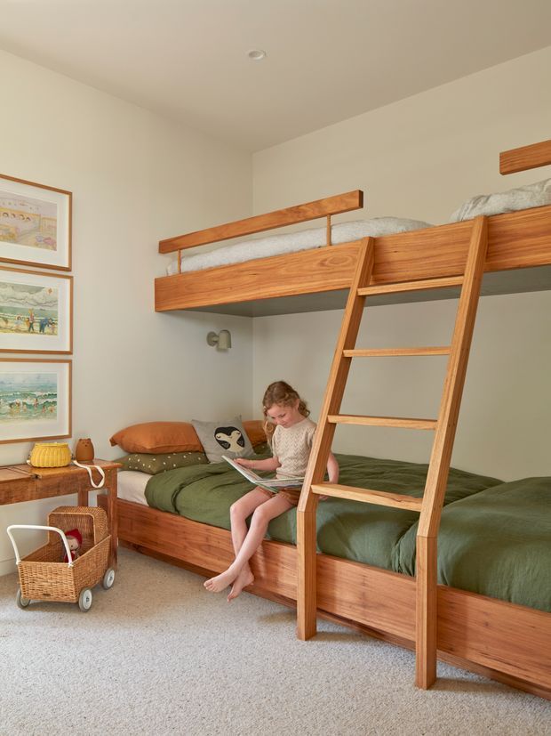 A warm, light-filled children's bedroom featuring a timber bunk-style loft bed with an integrated lower bed and ladder. The room has pale walls, soft carpet, green bedding, framed artwork, a small timber side table, and a woven toy trolley. A child is seated reading on the lower bed.