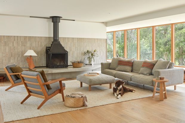 Bright, inviting living room with a large grey sectional sofa, timber-framed lounge chairs, a wood-burning fireplace with a stone or tile surround, a low upholstered ottoman, and large timber-framed windows overlooking trees. The room has pale timber flooring, a light neutral rug, and a warm, relaxed contemporary-natural aesthetic.