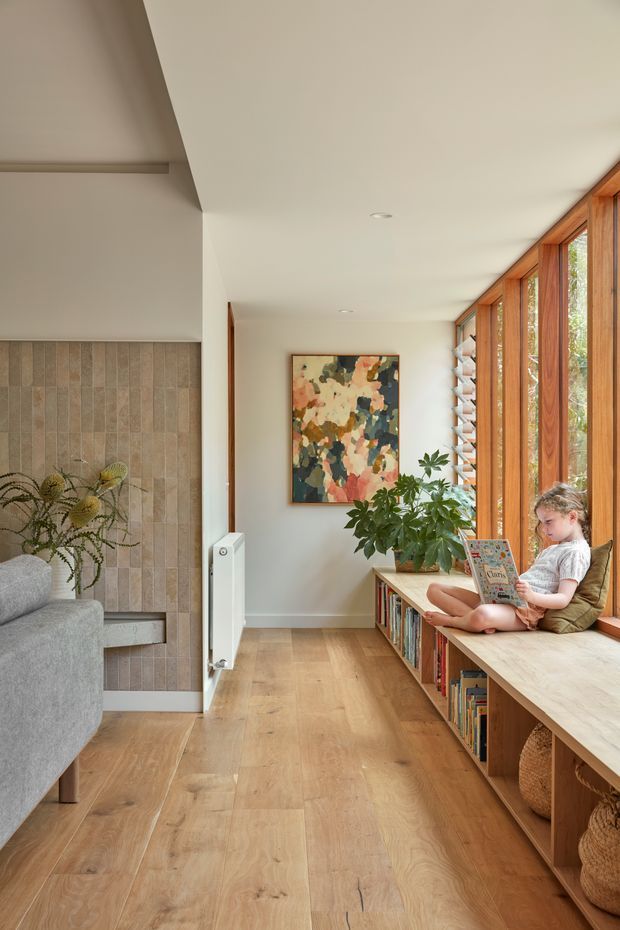 A bright, narrow residential living area with a full-height timber-framed window wall and integrated timber window seat/bookcase. The space features pale timber flooring, a white ceiling and walls, a vertically tiled feature wall, a grey sofa, indoor planting, and colourful abstract artwork. A child is seated reading on the built-in bench. The overall design is warm, contemporary, and connected to natural light and greenery outside.